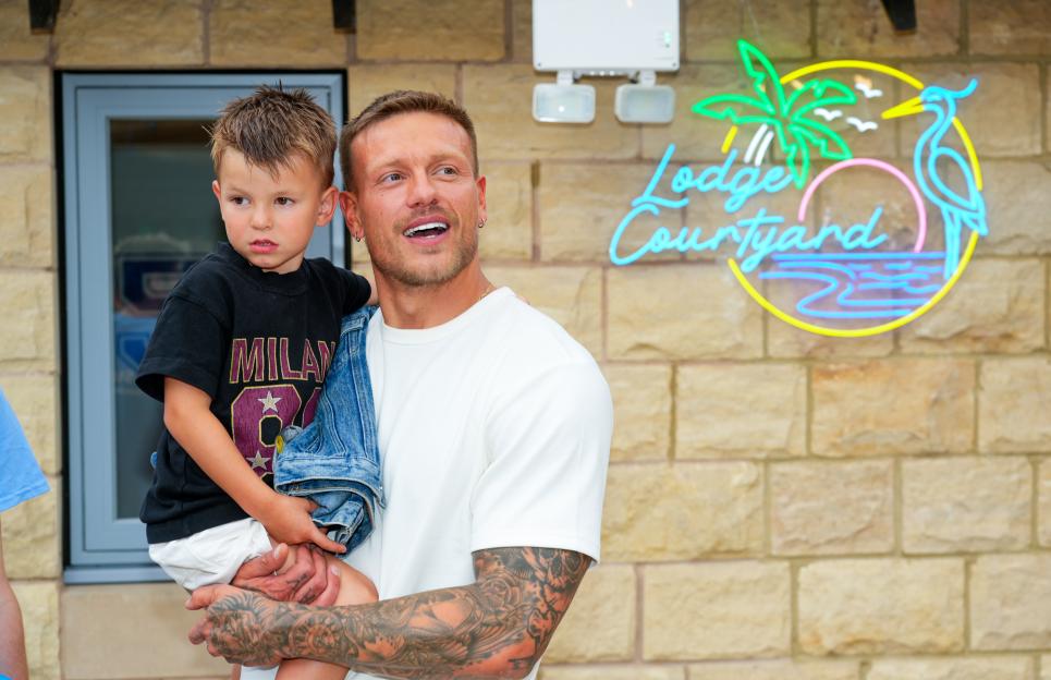 Alex Bowen holding a child in front of a brick wall with a neon sign that reads "Lodge Courtyard."