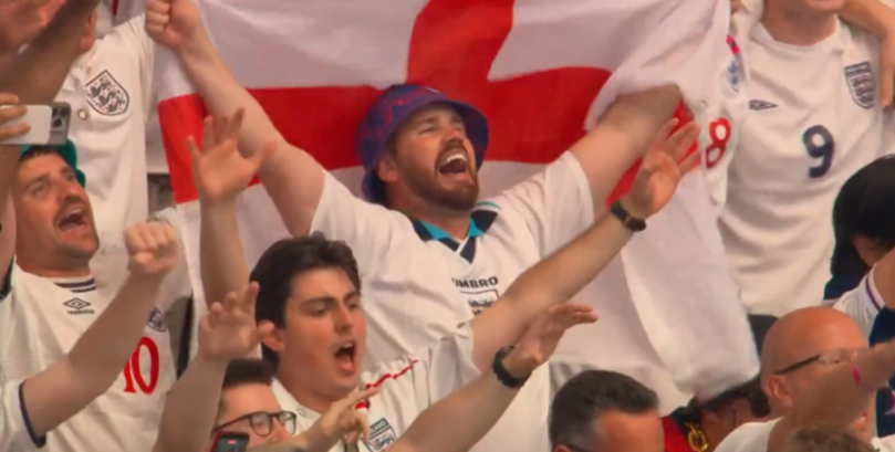 Fans celebrate while holding an England flag at a football match.
