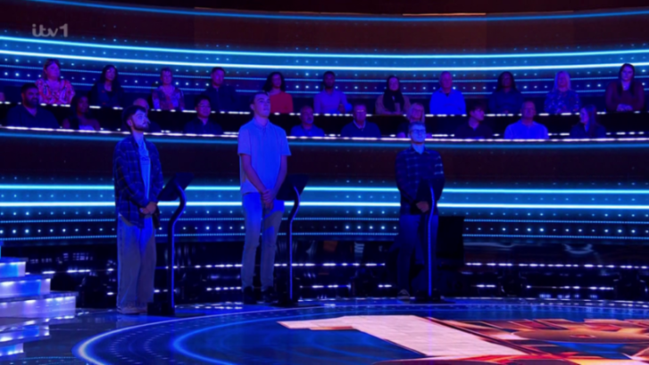 Three contestants stand in front of an audience during a game show illuminated by blue lights.