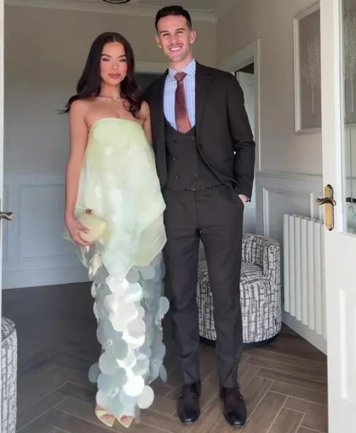 A smiling couple in formal attire standing in a home entryway, the woman in a strapless light green dress with large sequins, and the man in a dark brown suit with a striped shirt and reddish-brown tie.