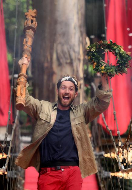 Sam Thompson raises a carved scepter and floral wreath above his head, celebrating his win as King of the Jungle.