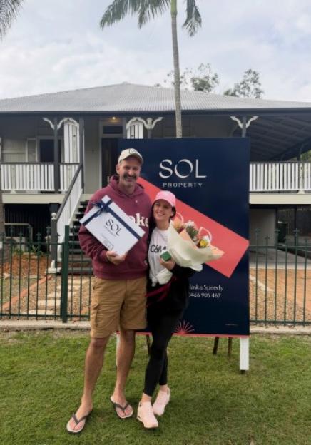 A man and a woman stand smiling in front of a house and a "SOL PROPERTY" sign, holding gifts.