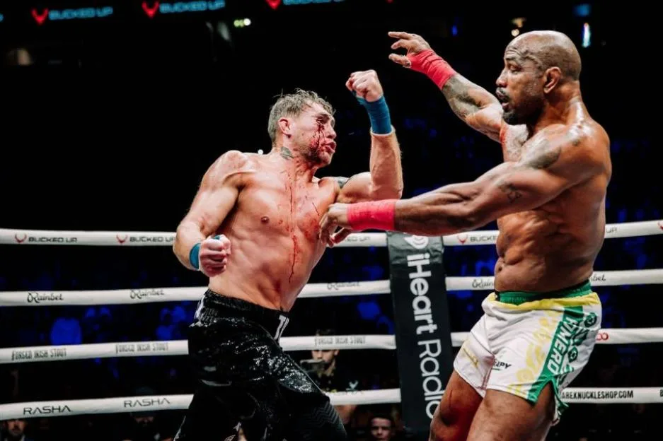 Two male boxers, one bleeding from the face, face each other in a boxing ring.