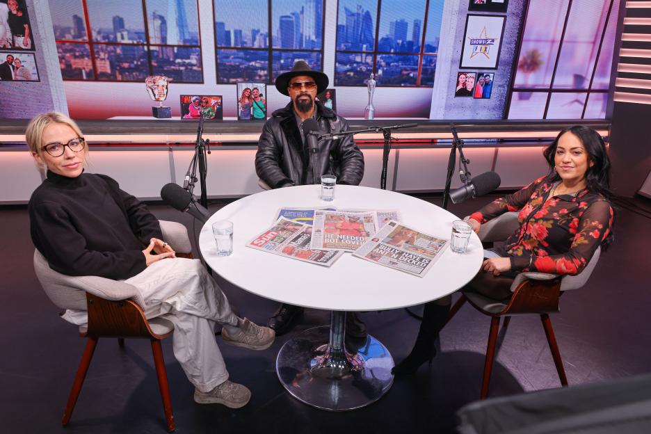 Three people sitting around a round table with newspapers spread out, microphones, and glasses of water.