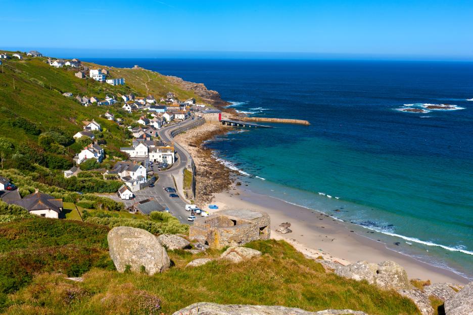 View over Sennen Cove on the Penwith peninsula, with houses on green hills, a road, beach, and blue sea.