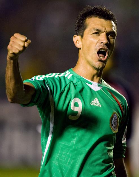 A Mexican soccer player in a green jersey with the number 9 and an Adidas logo clenches his fist and shouts in celebration.