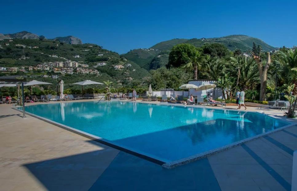 Outdoor swimming pool with people relaxing on lounge chairs and mountains in the background.