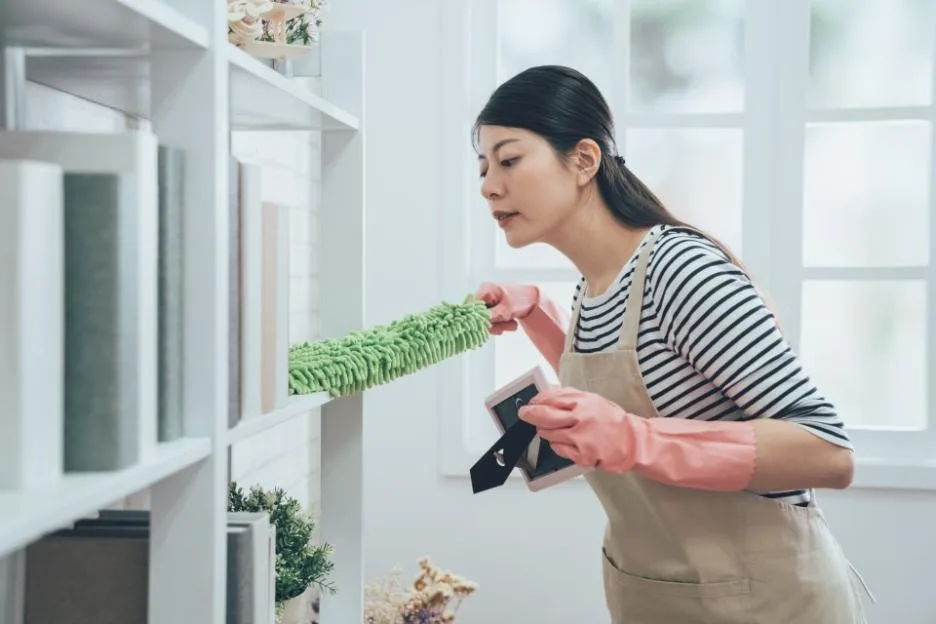 An Asian housekeeper wearing an apron and pink gloves dusts a bookshelf with a feather duster while carefully holding a picture frame.