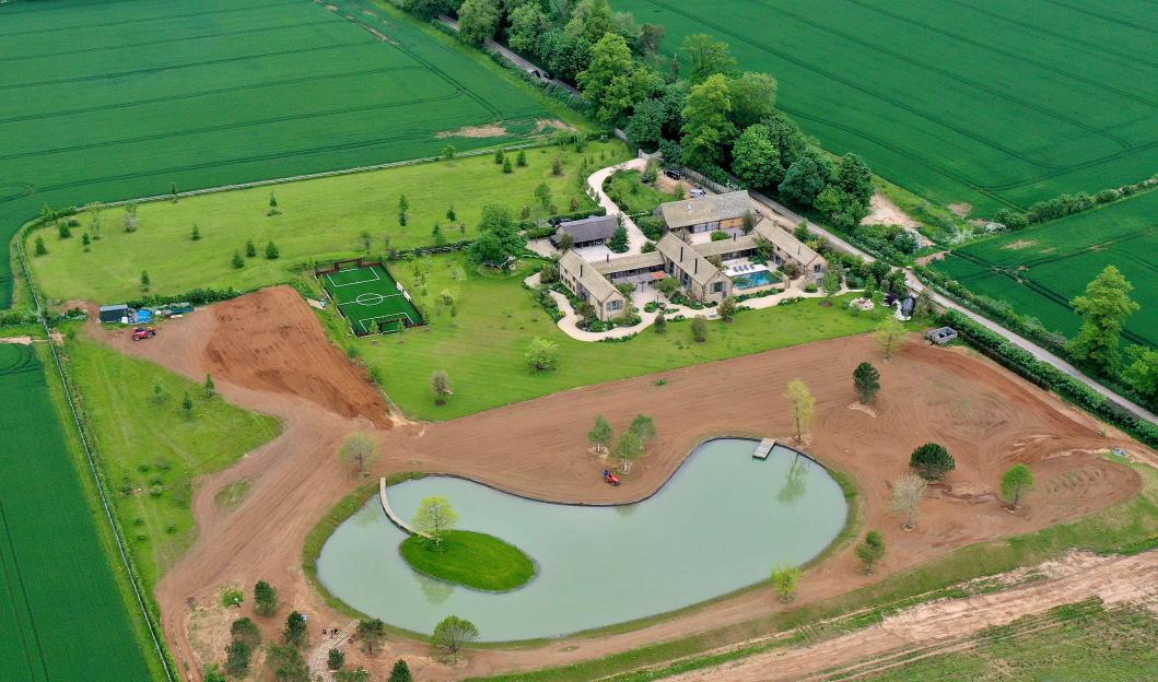 Aerial view of David and Victoria Beckham's home in Great Tew, featuring a large estate, a soccer field, and a pond with an island.