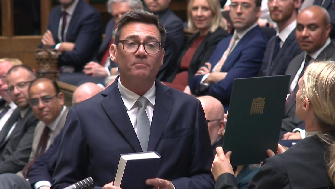 Andy Burnham being sworn in as an MP, holding a book, with another person holding a document with the UK Parliament crest.