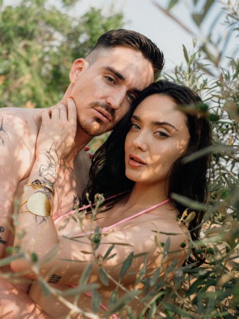A couple, a man and a woman, standing closely together among green leaves, both looking forward.