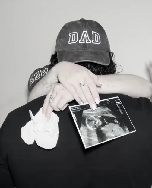 Lydia Rodford and her partner holding baby booties and a baby scan, with him wearing a 'Dad' hat and her wearing a 'Mum' top.