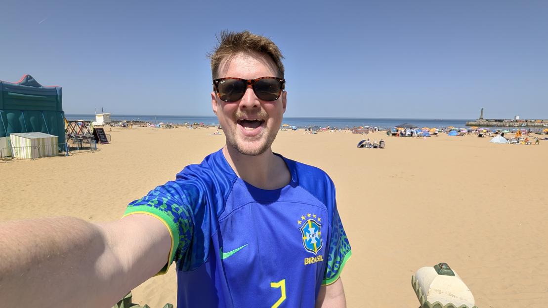 A man in a Brazil soccer jersey and sunglasses smiles at the camera on a sandy beach filled with people, umbrellas, and the sea in the background.