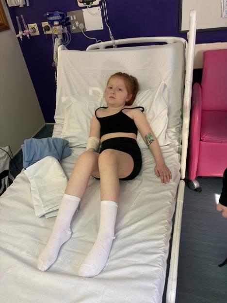 A red-headed girl in a hospital bed with a bandage on her left arm and a Pac-Man temporary tattoo on her right arm.