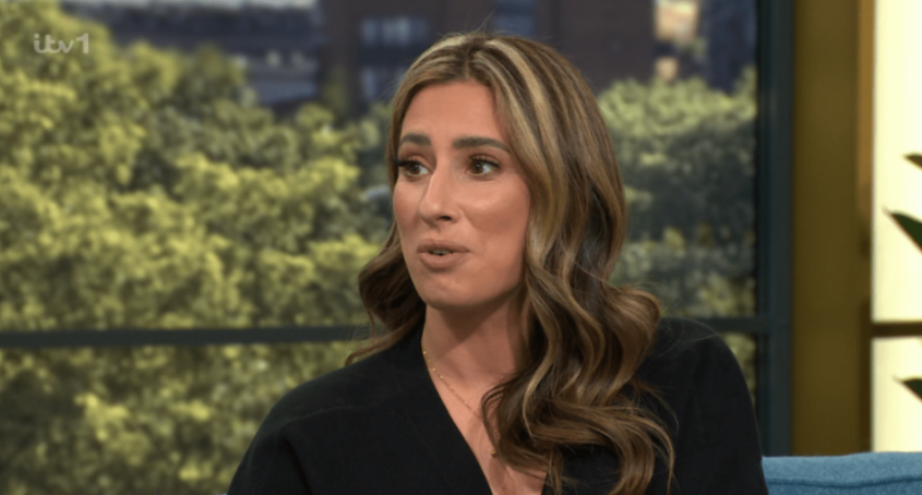 A woman with long wavy brown hair and highlights, wearing a black top and a gold necklace, speaks to the right.