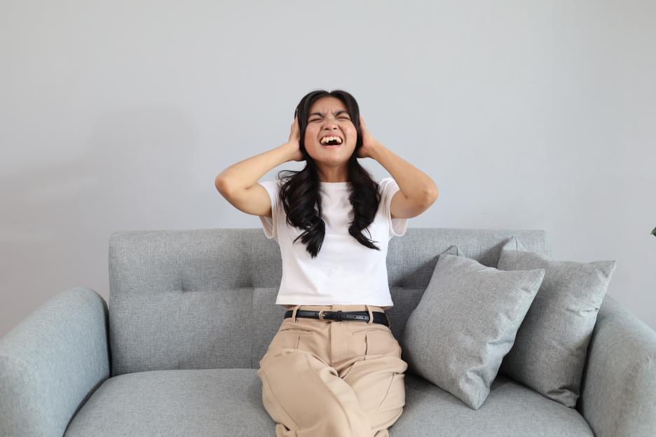 A young woman sitting on a gray sofa, shouting and covering her ears in frustration.