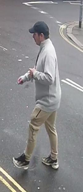 A person in a gray sweatshirt, tan pants, and black baseball cap walks across a paved surface.