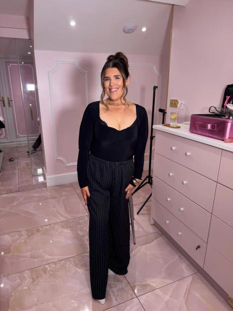 A smiling woman in a black top and pinstriped pants stands in a pink room.