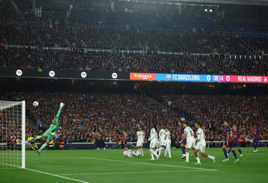 A soccer player in a blue and red striped jersey kicking the ball, with another player in a white jersey watching. The score shows BAR 1, RMA 0.