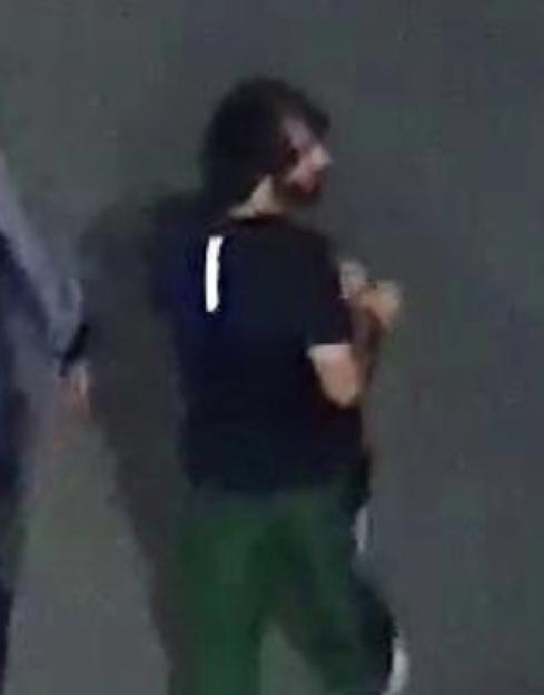 A man with dark hair and a beard, wearing a black shirt with a white vertical stripe on the back and dark green pants.