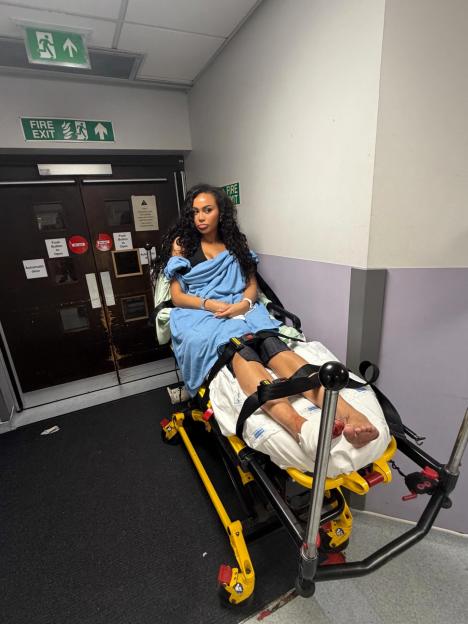 Ella Thomas, from Love Island, sitting on a hospital stretcher with her legs restrained and a blue blanket wrapped around her, looking straight ahead.