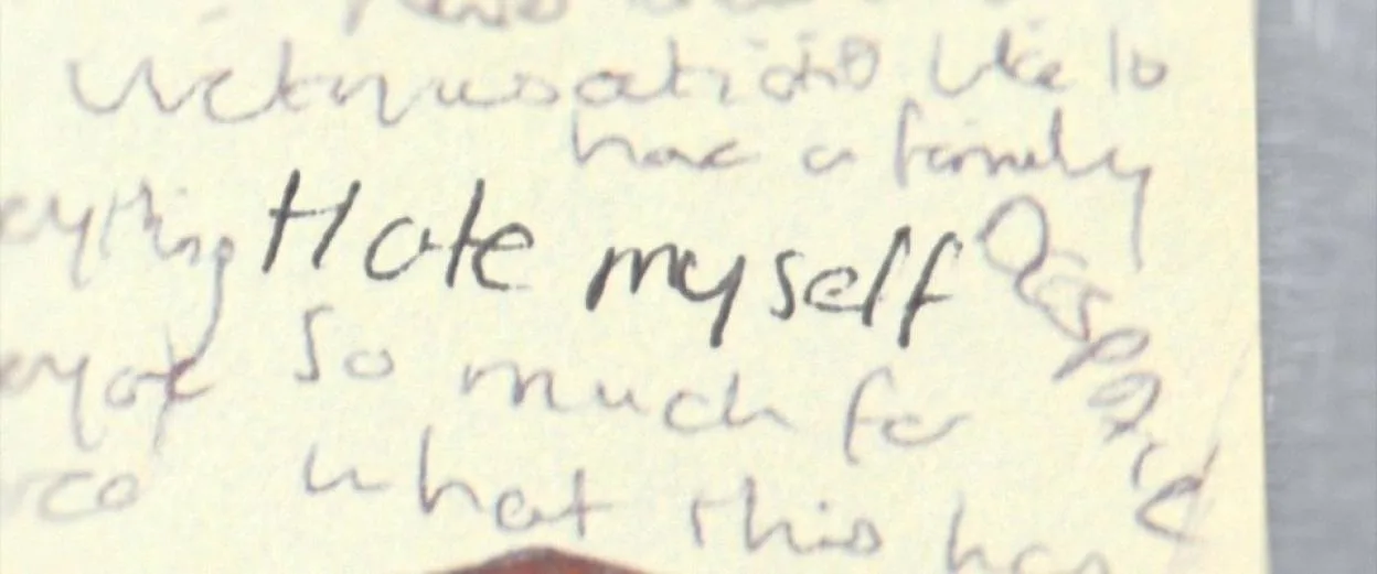 Handwritten text on paper stating "Hate myself" amidst other scribbled words.