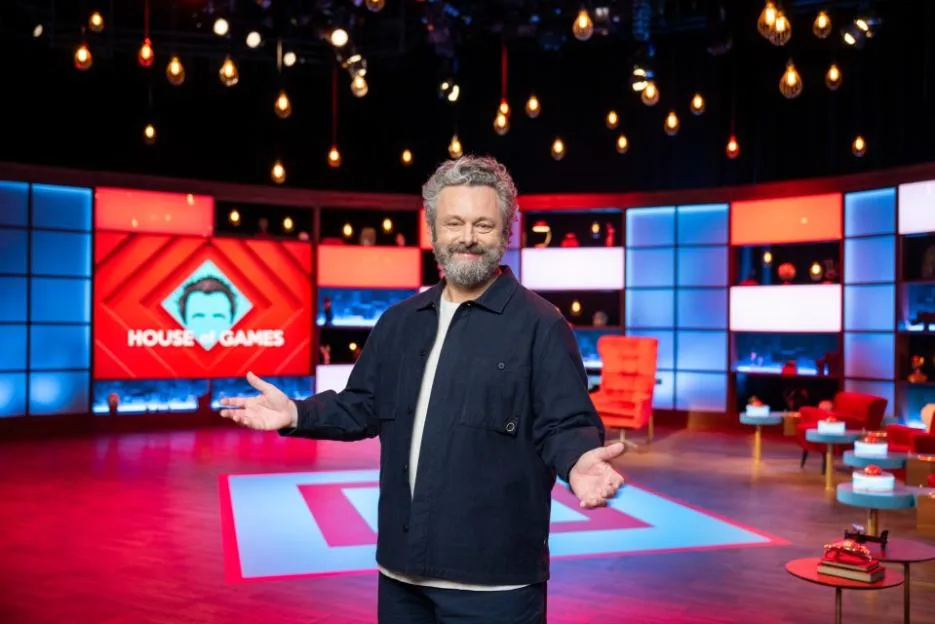 Michael Sheen stands on the set of House of Games, gesturing with his hands outstretched.
