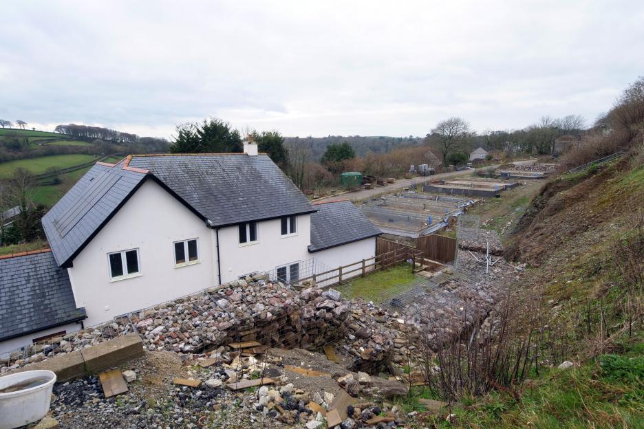 An abandoned construction site with a completed white house with solar panels, multiple house foundations, and gabion retaining walls.