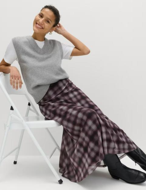 A woman wearing a gray sweater vest over a white t-shirt, a checked midi A-line skirt, and black boots.