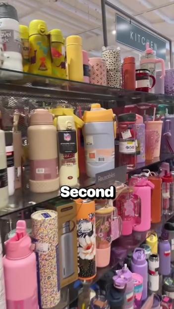A store shelf displaying a variety of colorful reusable water bottles and mugs, with a "KITCHEN" sign in the background.