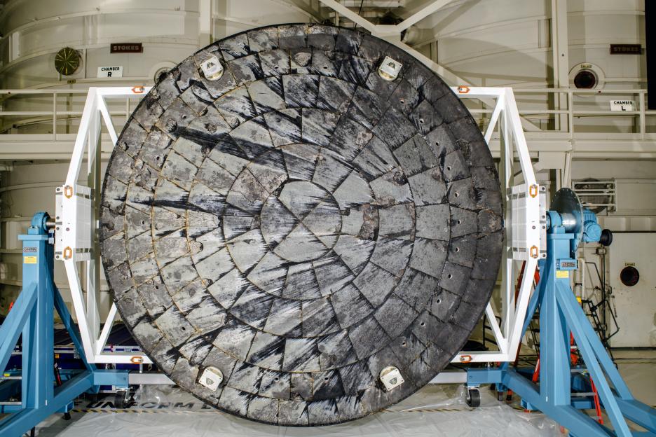 The Orion spacecraft's heat shield, covered in black and grey thermal protection tiles, on a blue and white stand inside a facility.