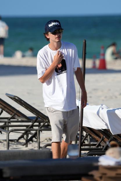 Timothée Chalamet standing on a beach, wearing a navy Nahmias hat, sunglasses, white Supreme x Wu-Tang Clan RZA T-shirt, and beige shorts.