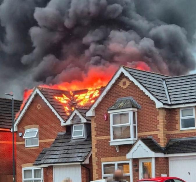 A house roof is engulfed in flames with large plumes of black smoke rising into the sky.