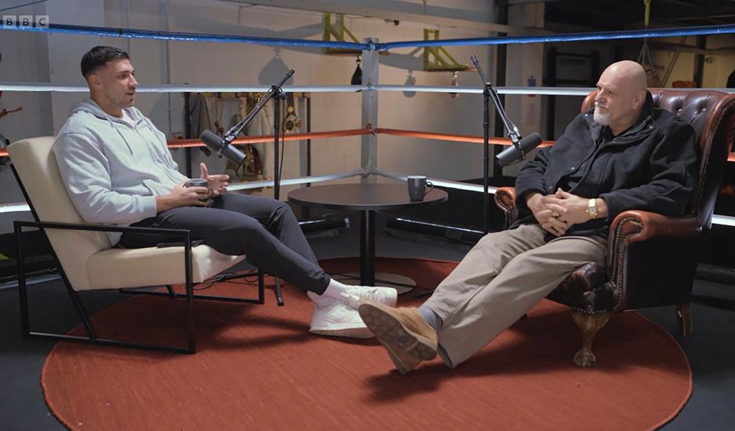 Tommy Fury and another man are seated in a boxing ring for a podcast, microphones visible between them.