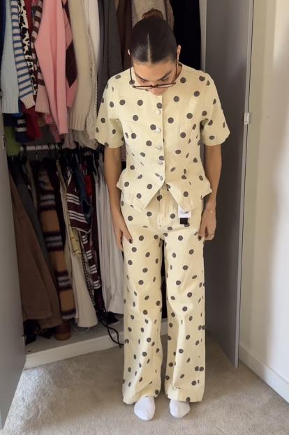 A woman wearing a cream-colored polka-dotted top and matching trousers stands in front of an open wardrobe.