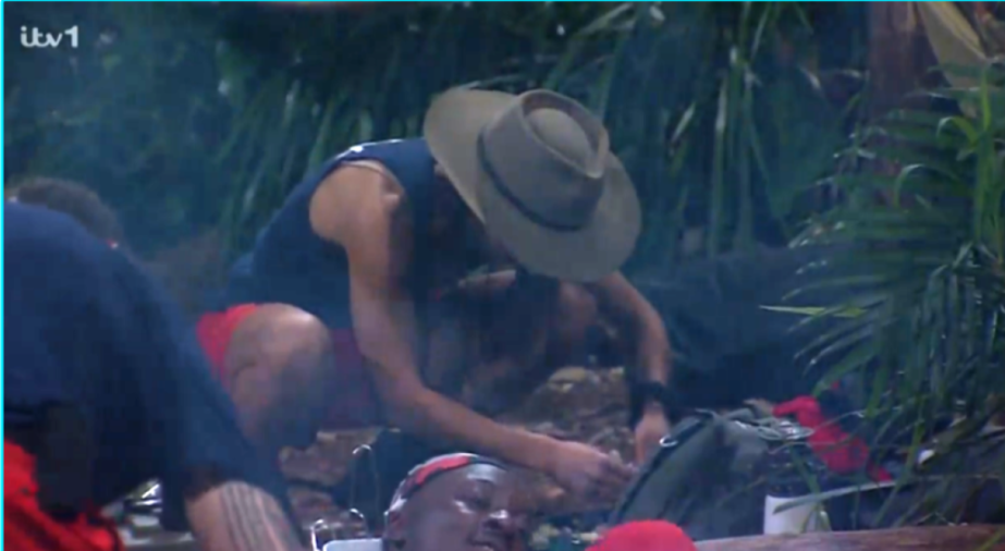 A man in a hat bending over to tend to something on the ground, surrounded by foliage.