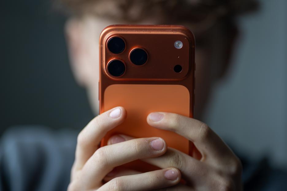 A close-up of a boy holding an iPhone in an orange case, with his face obscured by the phone.