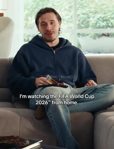 Man in a navy hoodie and jeans sitting on a sofa, holding a remote and food packaging, with text "I'm watching the FIFA World Cup 2026™ from home."