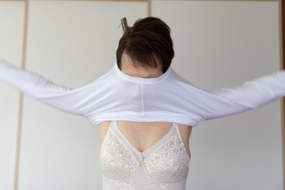 A senior woman pulling a white long-sleeved shirt over her head, revealing a lace bra.