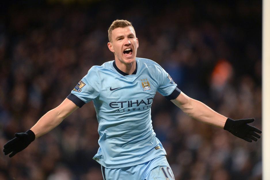 Soccer player Edin Dzeko in a light blue Manchester City kit with his arms outstretched, celebrating a goal during a match against Newcastle United.