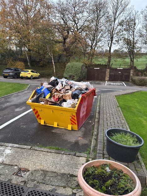 A large yellow and red skip full of trash sitting on an asphalt driveway next to a grassy area.