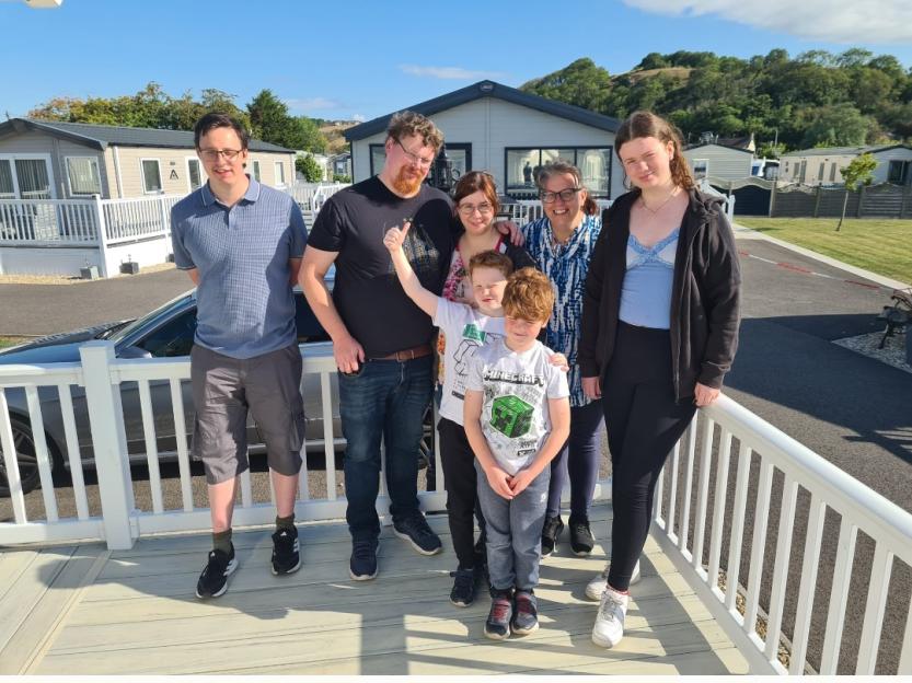 A family of seven, consisting of four adults and three children, stands on a wooden deck in front of mobile homes.