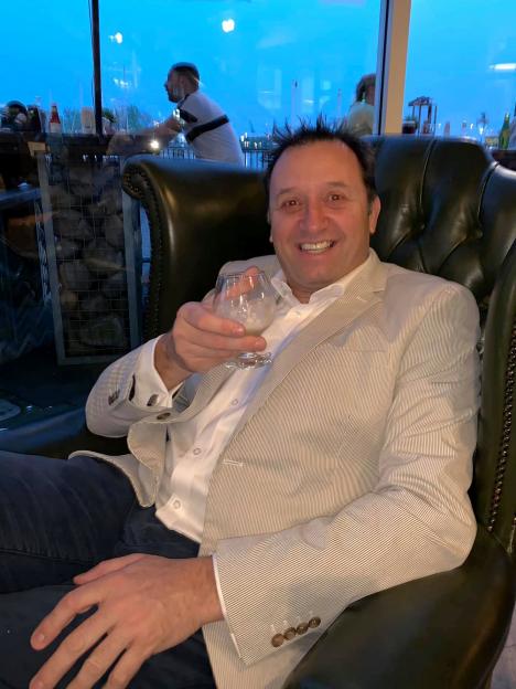 Gareth Lloyd, a man with spiky dark hair, wearing a white shirt and pinstriped jacket, smiles while holding a drink.