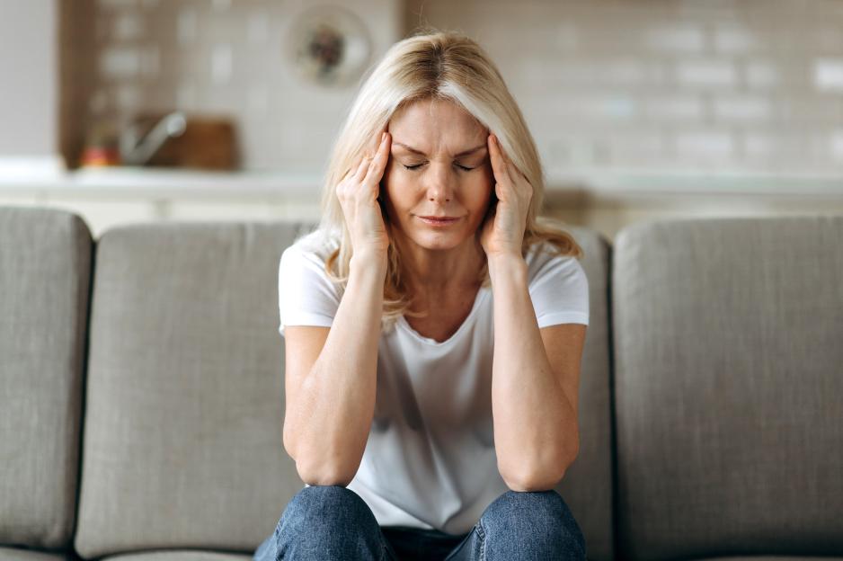 A blonde woman sits on a couch, holding her head in her hands due to a headache or distress.