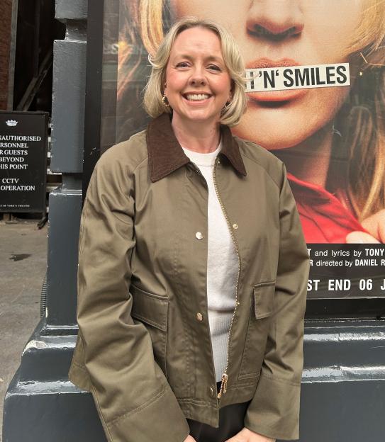 A smiling woman stands in front of a play poster featuring a large image of actress Rebecca Lucy Taylor.