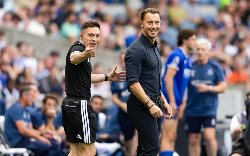 Newcastle Head Coach Matthias Jaissle smiling at the camera as a fourth official speaks to him on the sidelines.
