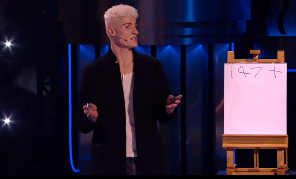 A man with bleached hair stands next to a whiteboard with "147+" written on it.
