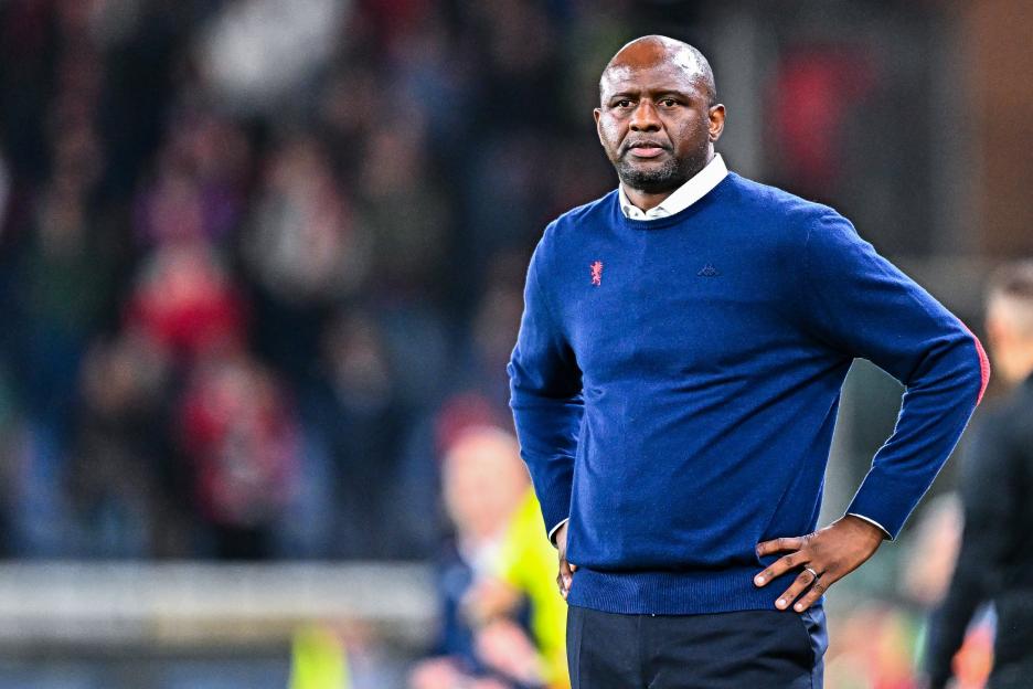 A dark-skinned man with a short beard and bald head wears a blue sweater over a white collared shirt, hands on his hips.
