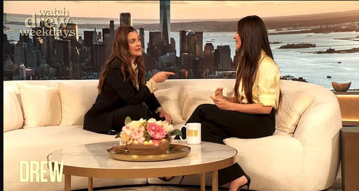 Drew Barrymore and Olivia Munn sitting on a couch on "The Drew Barrymore Show" set.