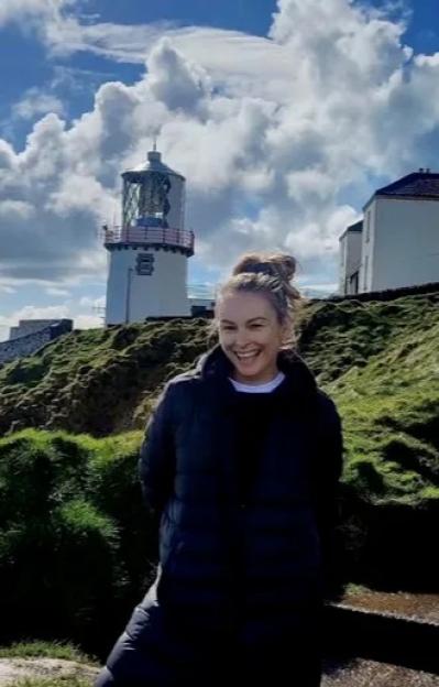 A woman with long blonde hair and a white feathered jacket laughs with her hand on her chin.